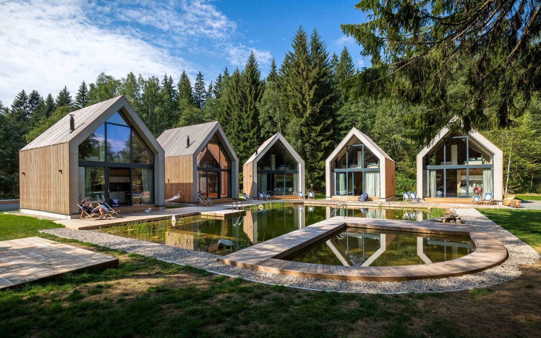 Rząd domków nad stawem, za nimi świerkowy las